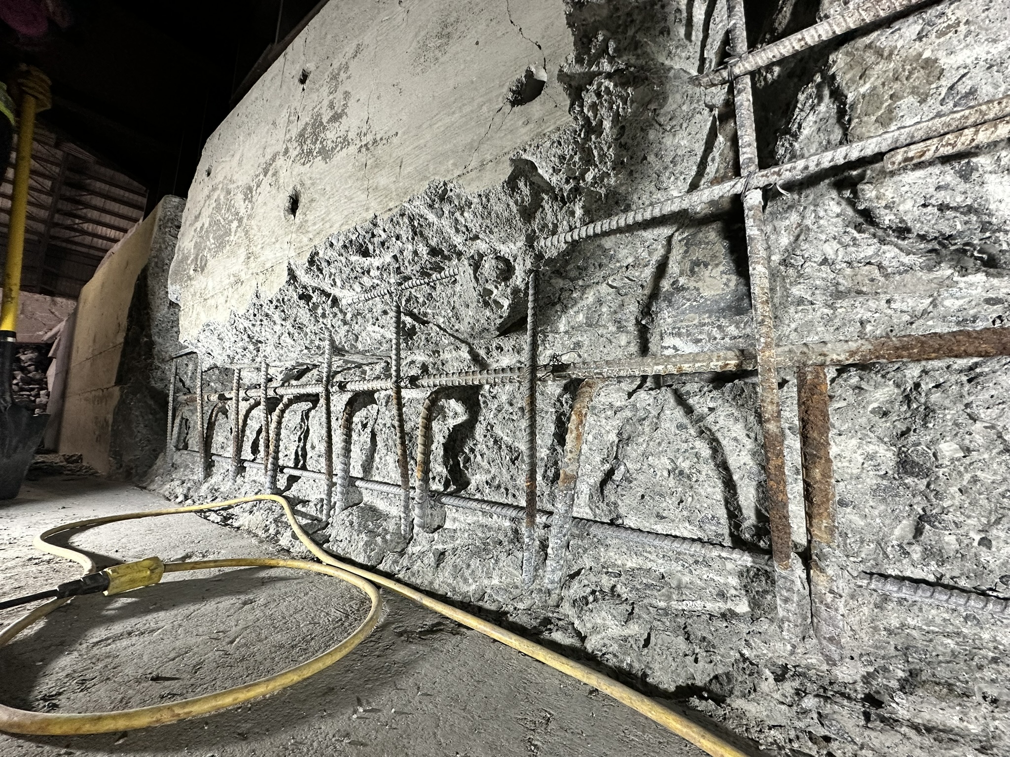 Concrete Repair Services Jacksonville, FL | Con Cure USA IMG 5957 Exposed concrete wall with rusted rebar, showing layers of rough texture; yellow hose lies on the dusty ground.