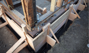 Construction repair site showing a wooden foundation frame with metal support bolts, ready for pouring concrete, on a dirt surface.